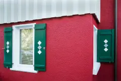Fassadendetail mit rotem Putz, weißer Holzverkleidung und grünen Fensterläden