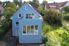 Hellblaues Einfamilienhaus mit Satteldach und kubischem Anbau. Vogelperspektive, frontal.